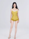 Full body shot of a woman in a mustard yellow one-piece swimsuit with floral trim and ruffled shorts, wearing white lace-up sandals.