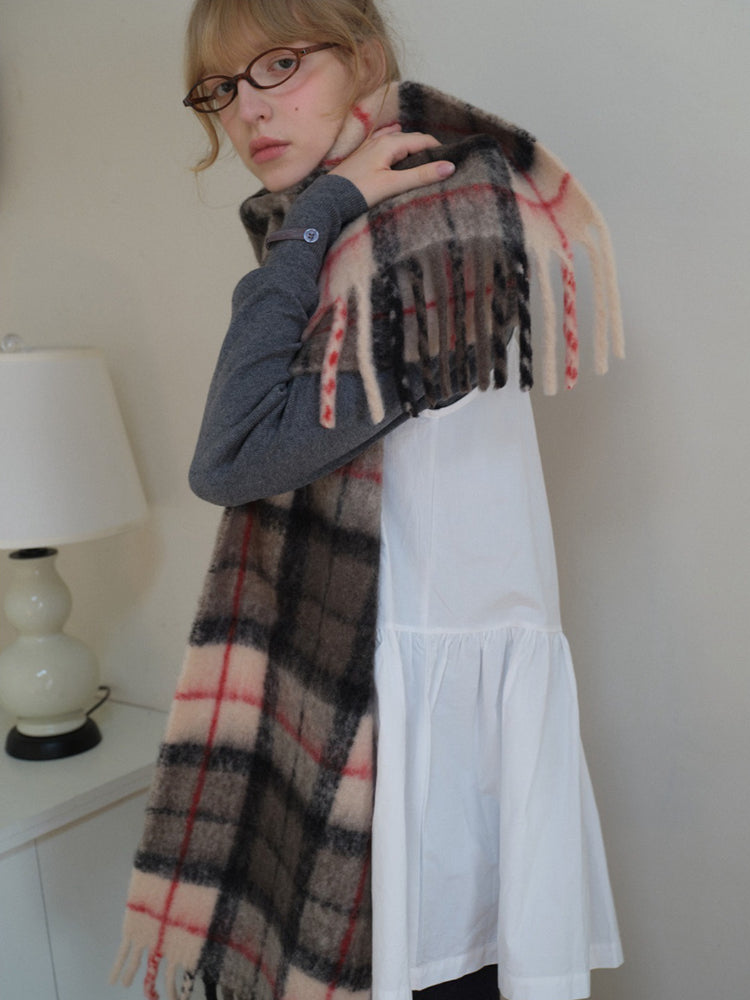 Model in a grey sweater and white dress, adjusting a large plaid scarf with fringe.