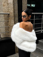 Woman wearing a cream faux fur cropped jacket and grey trousers, smiling outdoors.
