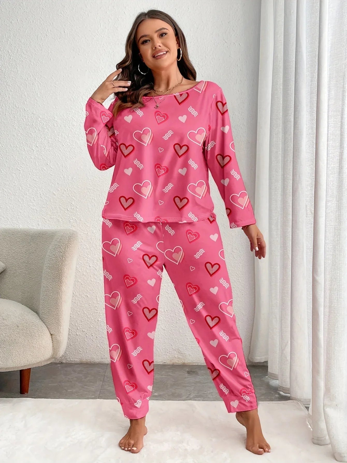 Woman wearing a pink long-sleeved pajama set with an all-over heart print, standing and posing. Main image