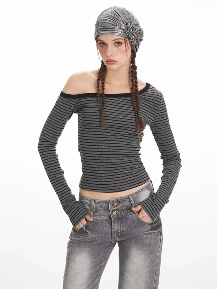 Front view of a woman wearing a grey and black striped off-the-shoulder long sleeve top and grey wash jeans, with hands in pockets.