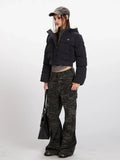 Side profile of a woman wearing a black cropped puffer jacket with a hood, grey t-shirt, and camouflage cargo pants, holding a black tote bag.