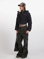 Side profile of a woman wearing a black cropped puffer jacket with a hood, grey t-shirt, and camouflage cargo pants, holding a black tote bag.