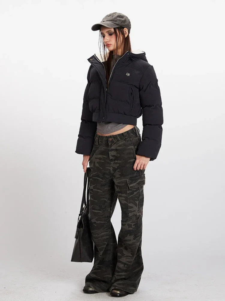 Side profile of a woman wearing a black cropped puffer jacket with a hood, grey t-shirt, and camouflage cargo pants, holding a black tote bag.