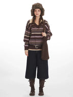 Woman wearing a brown striped knit sweater with a v-neck and collar detail, dark wash wide-leg cropped jeans, brown leather boots, a brown belt, and a brown faux fur hat with ear flaps.