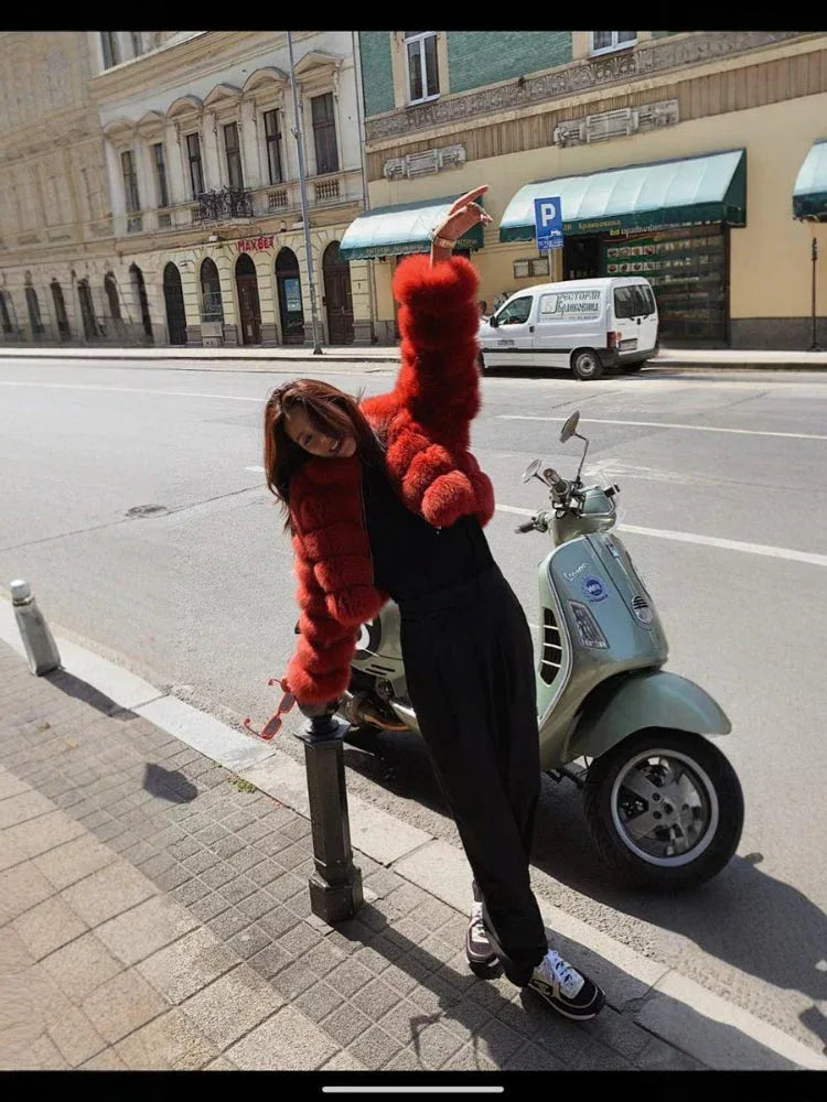 Woman wearing a red faux fur cropped jacket, black pants, and sneakers, posing next to a scooter.