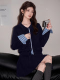 Close-up of a woman in a navy knit cardigan with blue gingham cuffs, holding a small silver camera.