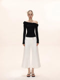 Full-body view of a woman wearing a black long-sleeved ruffled off-the-shoulder top and white wide-leg pants.