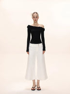 Full-body view of a woman wearing a black long-sleeved ruffled off-the-shoulder top and white wide-leg pants.