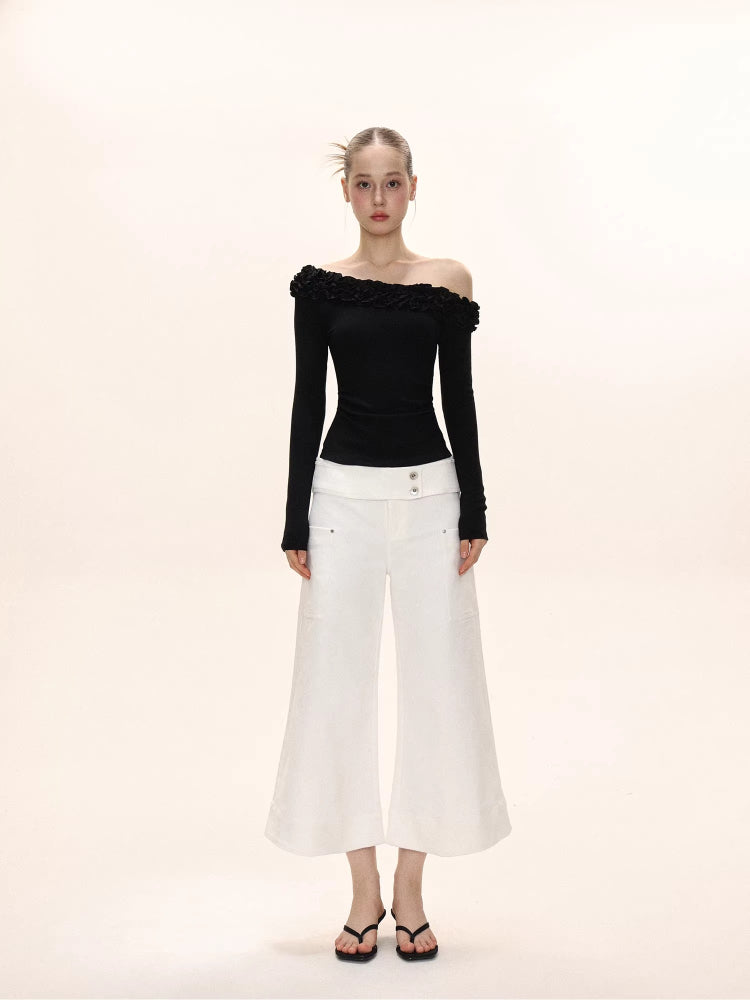Full-body view of a woman wearing a black long-sleeved ruffled off-the-shoulder top and white wide-leg pants.