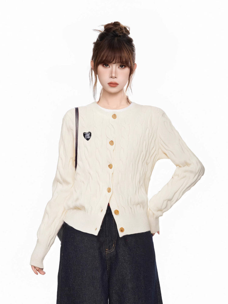 Woman wearing a cream cable knit cardigan with gold buttons and an embroidered heart detail on the left chest, paired with dark wash wide-leg jeans.