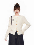 Front view of a cream cable knit cardigan with a crew neckline and long sleeves, featuring gold buttons and a black embroidered heart patch.