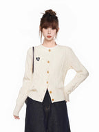 Front view of a cream cable knit cardigan with a crew neckline and long sleeves, featuring gold buttons and a black embroidered heart patch.