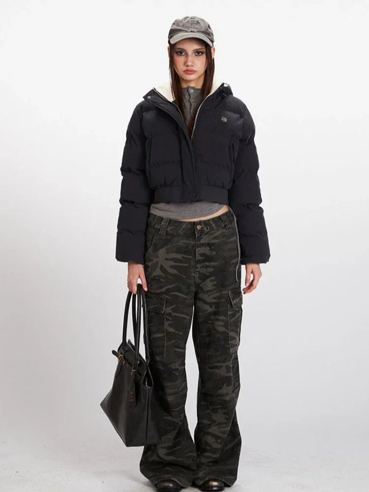 Woman wearing a black cropped puffer jacket with a hood, grey t-shirt, and camouflage cargo pants, holding a black tote bag.