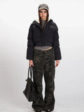 Woman wearing a black cropped puffer jacket with a hood, grey t-shirt, and camouflage cargo pants, holding a black tote bag.