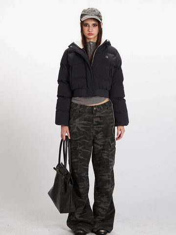 Woman wearing a black cropped puffer jacket with a hood, grey t-shirt, and camouflage cargo pants, holding a black tote bag.