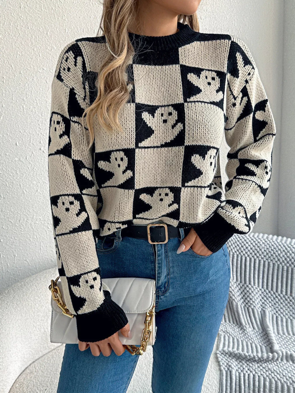 Woman wearing a black and cream checkerboard knit sweater with ghost print, paired with blue jeans and a white clutch.
