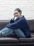 Woman sitting on a brown leather couch, hugging her knees, wearing a navy blue cable knit sweater and light wash jeans.