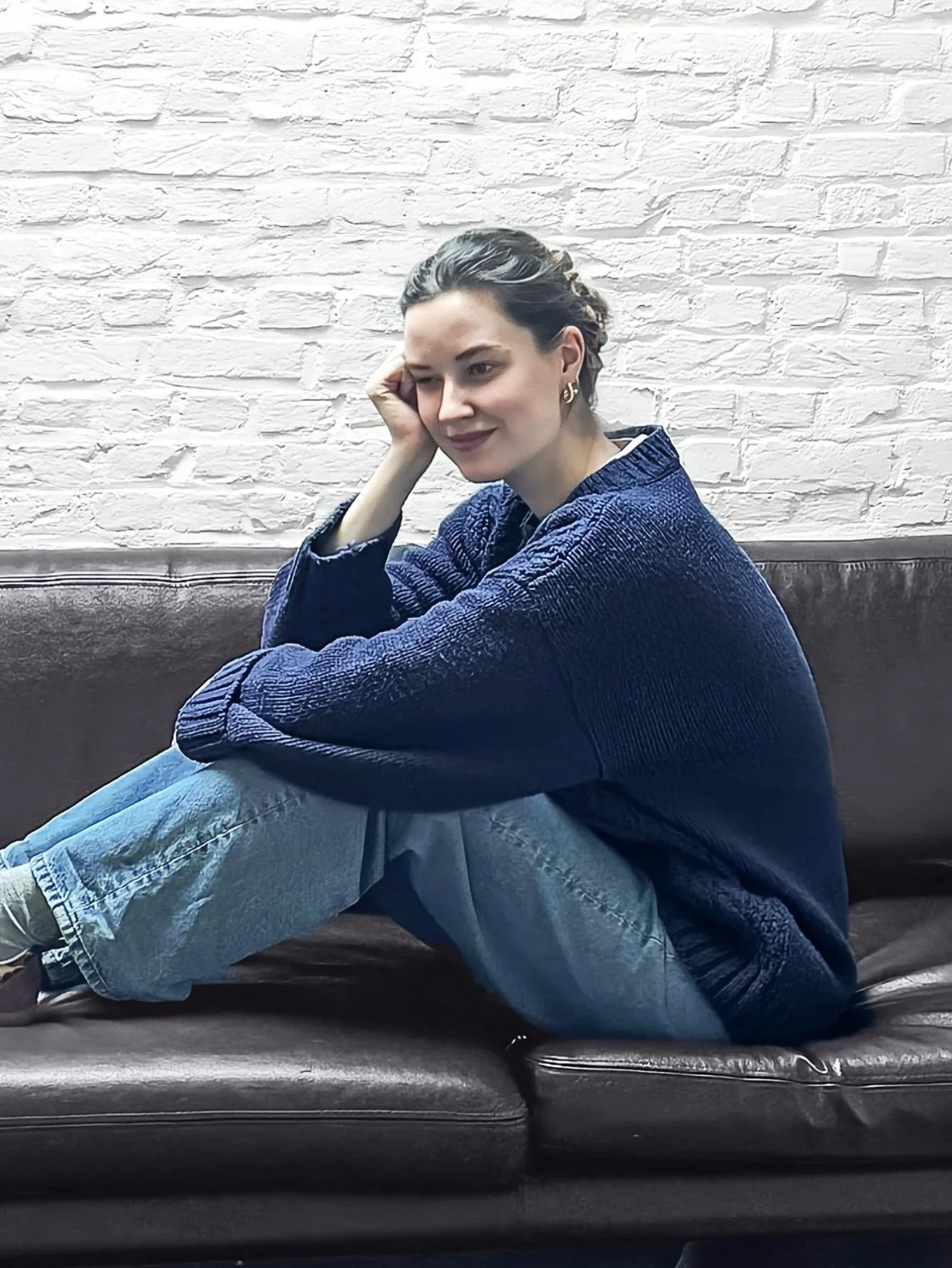 Woman sitting on a brown leather couch, hugging her knees, wearing a navy blue cable knit sweater and light wash jeans.