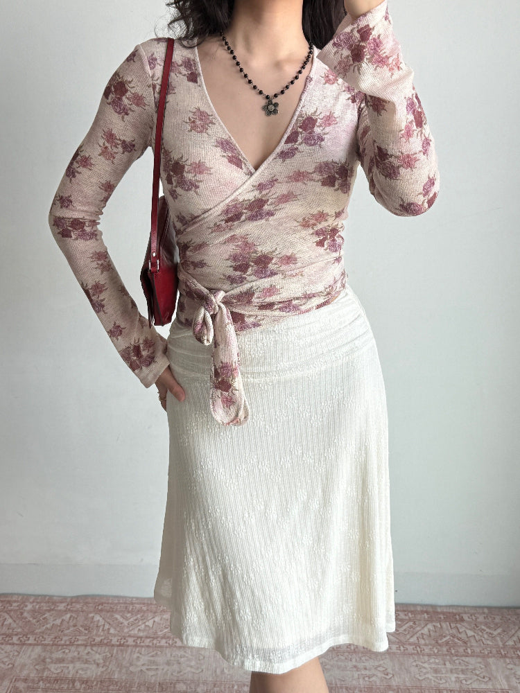 Woman wearing a floral wrap top, white skirt, and a red shoulder bag, posing against a white background.