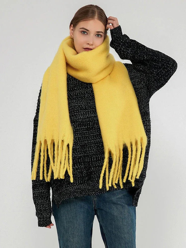 Woman wearing a bright yellow scarf with fringes around her neck, styled with a dark grey sweater and blue jeans.