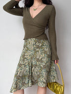 Full body shot of a woman in a green ribbed wrap top and a paisley print skirt, holding a yellow handbag.