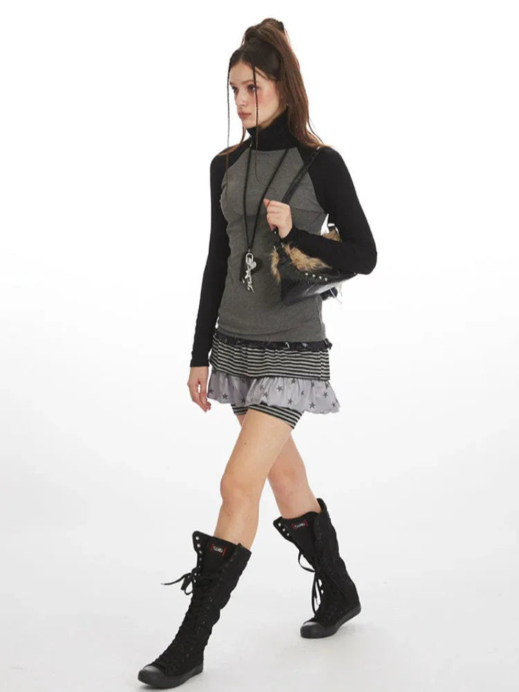 Woman walking forward wearing a grey long-sleeved turtleneck top with black raglan sleeves, layered grey and black striped shorts, and black lace-up boots.