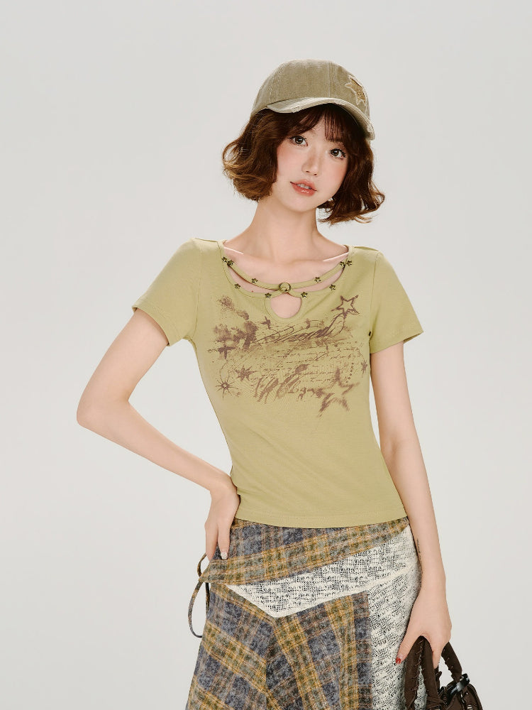 Woman wearing a light green short-sleeve fitted t-shirt with a cut-out neckline detail featuring star embellishments and a central ring. The t-shirt has a graphic print on the front and is paired with a plaid skirt.