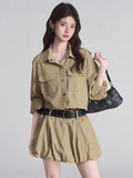 Woman wearing a khaki camp collar shirt dress with epaulet details and dual chest pockets, cinched at the waist with a black studded belt.