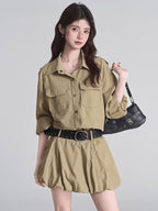 Woman wearing a khaki camp collar shirt dress with epaulet details and dual chest pockets, cinched at the waist with a black studded belt.