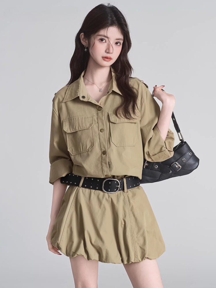 Woman wearing a khaki camp collar shirt dress with epaulet details and dual chest pockets, cinched at the waist with a black studded belt.