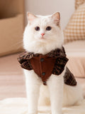 A white cat wearing a brown plaid pet dress with a ruffled collar and pleated skirt.