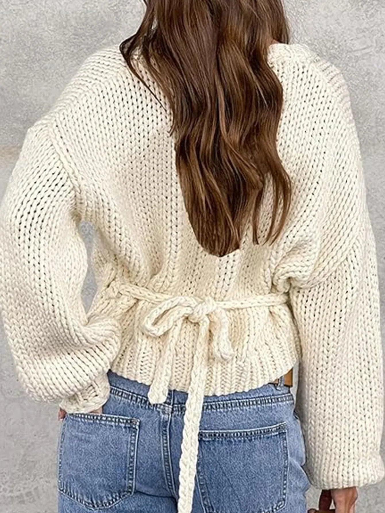 Back view of a woman in a cream chunky knit wrap sweater with a tie waist.