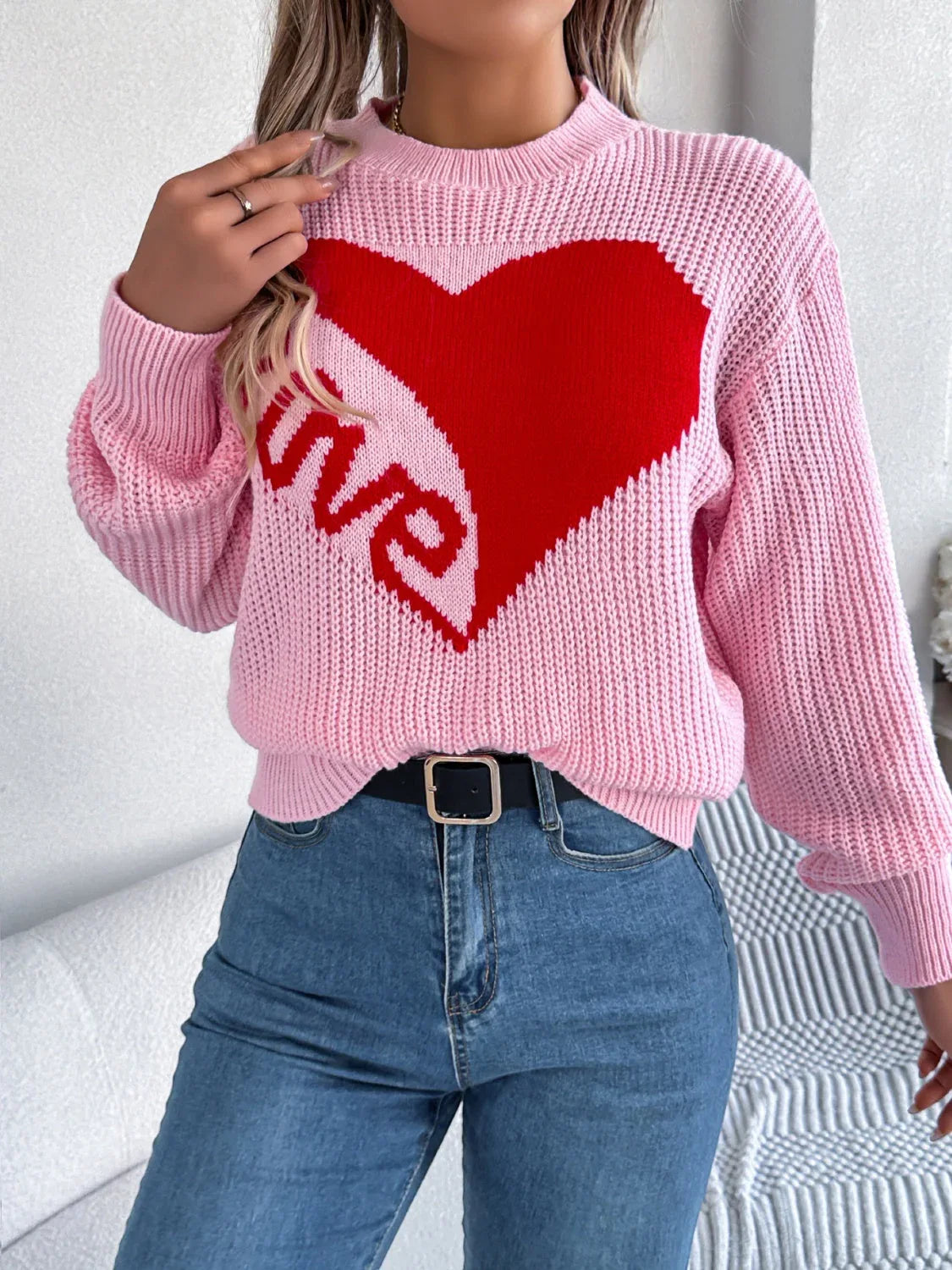 Woman styled in a pink knit sweater with a heart graphic, paired with blue jeans and a black belt.