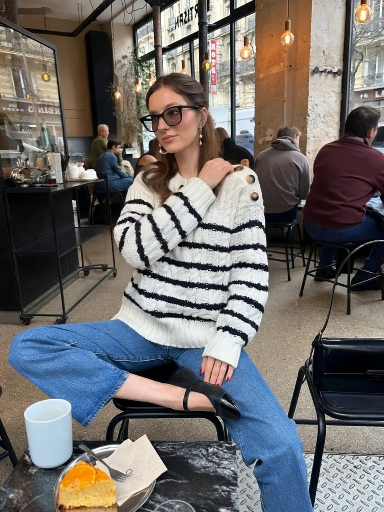 Woman in a white and black striped knit sweater with button details on the shoulder, blue denim jeans, and black ballet flats, posing in a cafe.
