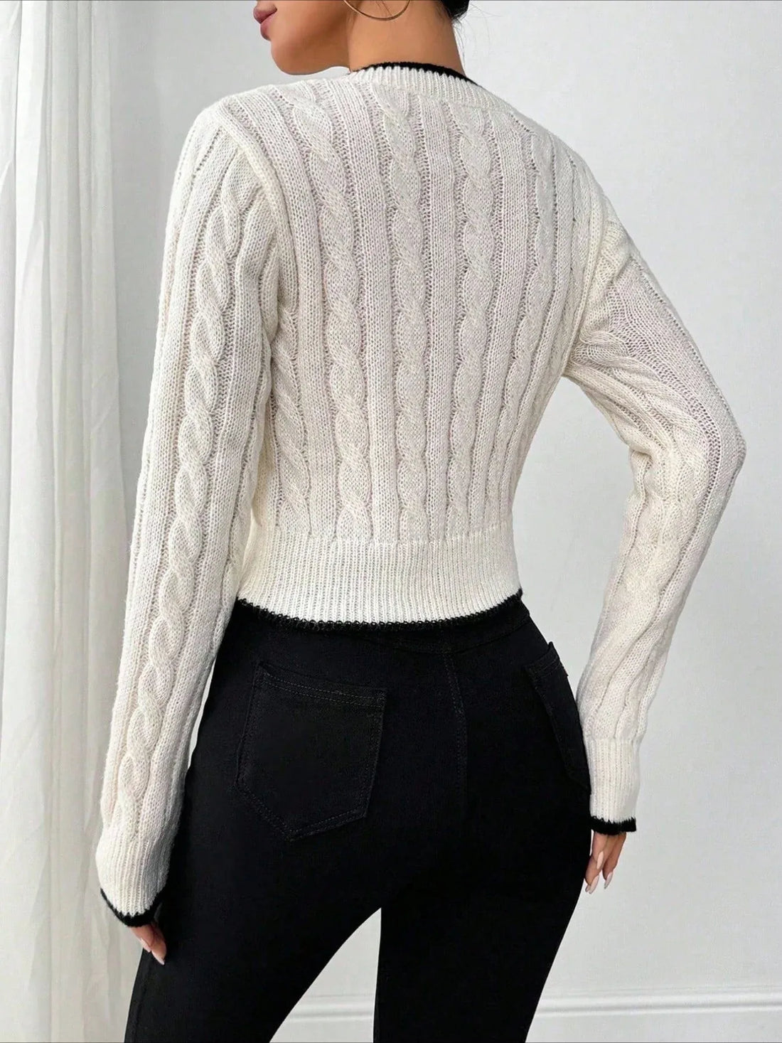 Woman wearing a white cable knit V-neck cropped sweater with black trim and an embroidered bow detail.
