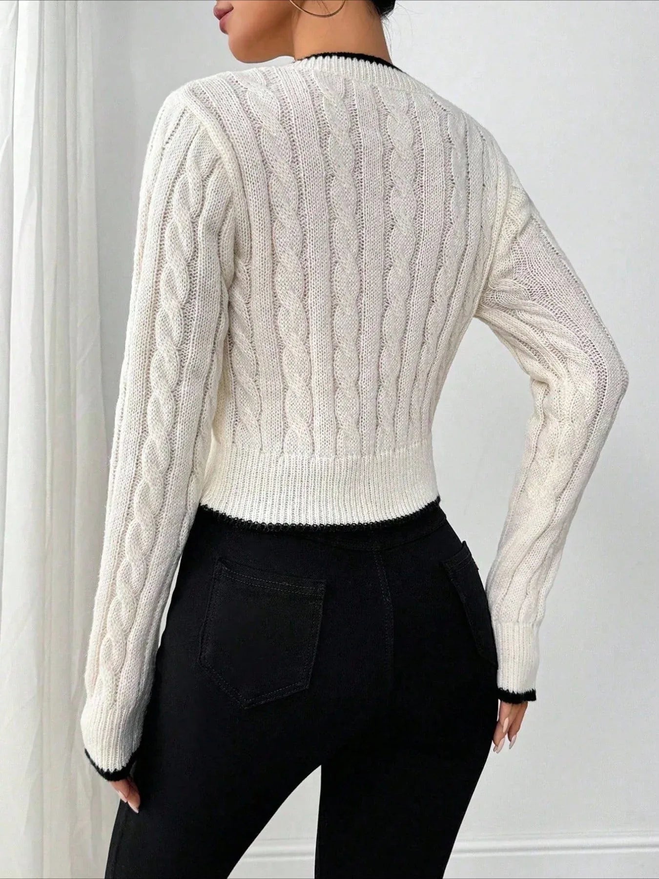 Back view of a woman wearing a white cable knit V-neck cropped sweater with black trim.