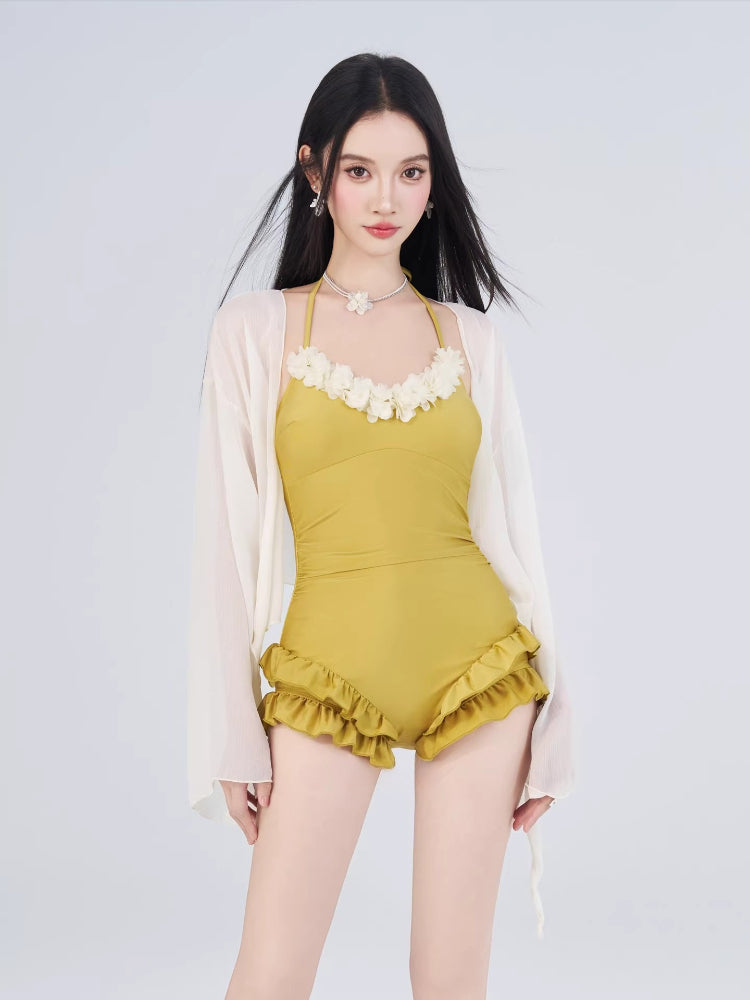 Woman wearing a mustard yellow one-piece swimsuit with floral trim and ruffled shorts, and a sheer white cover-up, standing against a white background.