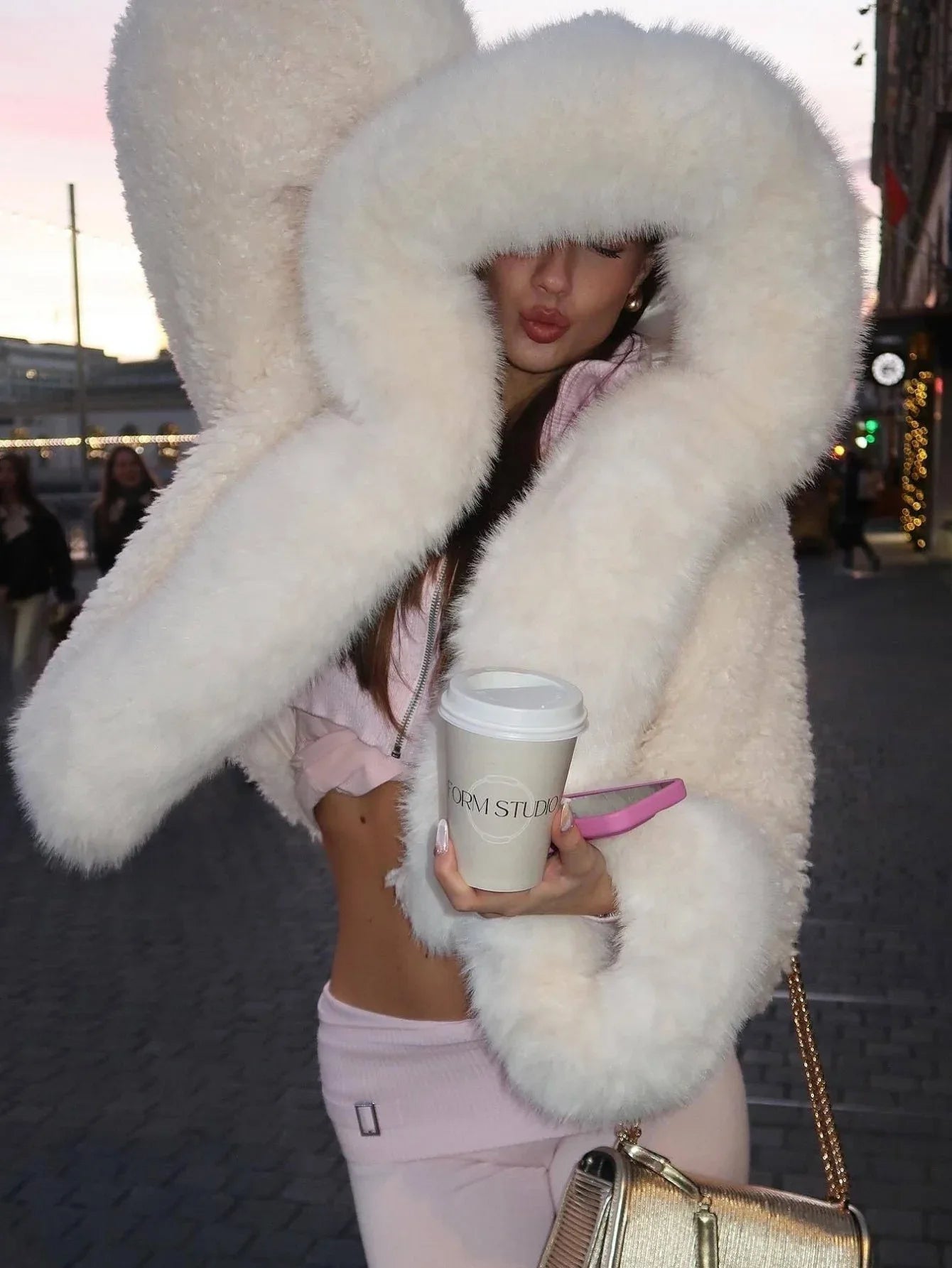 Woman wearing a cream-colored faux fur cropped jacket with an oversized hood and holding a coffee cup Main image
