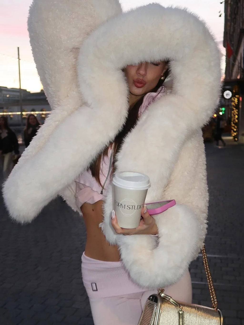 Woman wearing a cream-colored faux fur cropped jacket with an oversized hood and holding a coffee cup