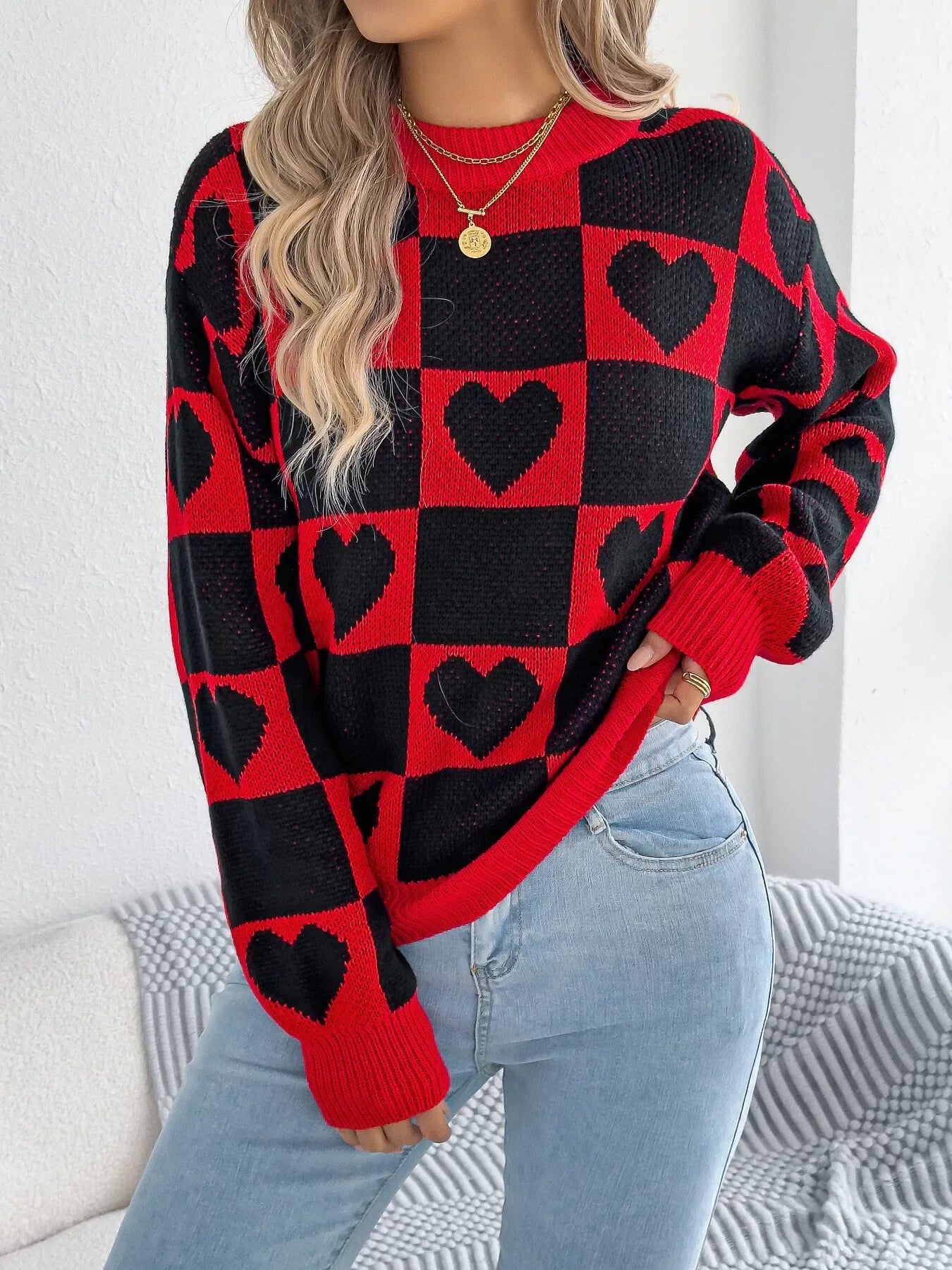 Close-up of a woman wearing a red and black checkerboard knit sweater with black heart patterns and light wash denim jeans.