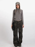 Model wearing a grey long-sleeve mock neck cropped top and camouflage cargo pants, accessorized with a grey baseball cap and a black fringed crossbody bag.