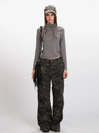 Model wearing a grey long-sleeve mock neck cropped top and camouflage cargo pants, accessorized with a grey baseball cap and a black fringed crossbody bag.