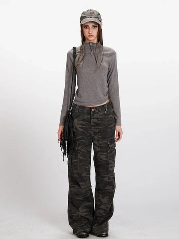 Model wearing a grey long-sleeve mock neck cropped top and camouflage cargo pants, accessorized with a grey baseball cap and a black fringed crossbody bag.