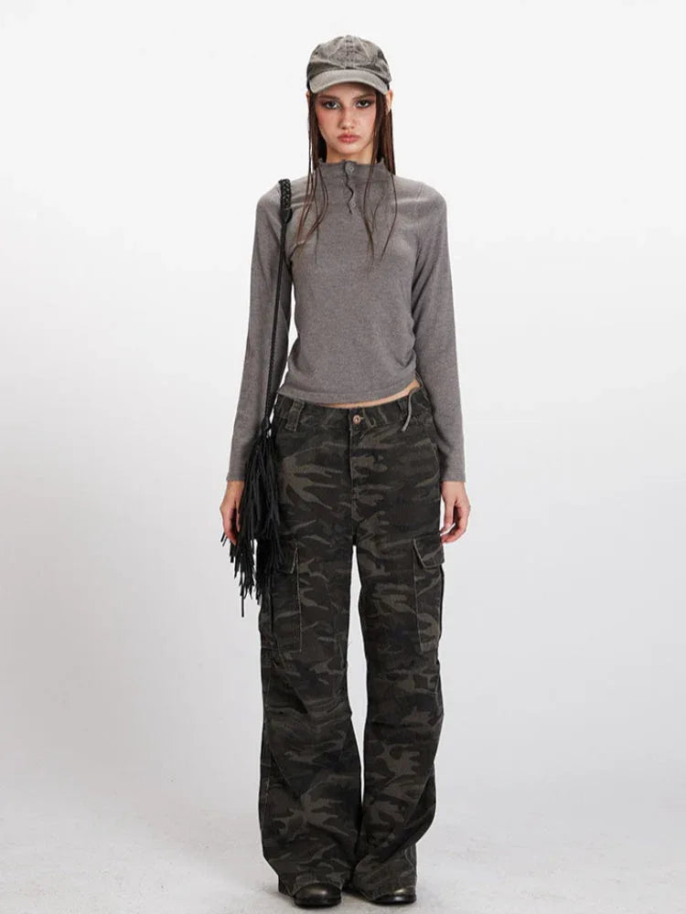 Model wearing a grey long-sleeve mock neck cropped top and camouflage cargo pants, accessorized with a grey baseball cap and a black fringed crossbody bag.