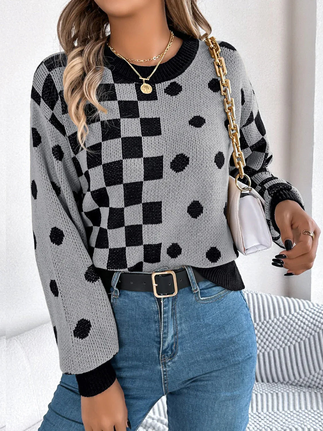 Woman holding a white handbag and wearing a grey knit sweater with black checkerboard and polka dot patterns, and blue jeans.