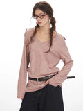 Close-up front view of a woman wearing a dusty rose long-sleeve knit top with a V-neck and chest pocket. She has braids and glasses.