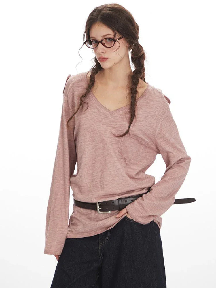 Close-up front view of a woman wearing a dusty rose long-sleeve knit top with a V-neck and chest pocket. She has braids and glasses.
