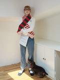 Model wearing a white sweater with an embroidered initial, blue jeans, and a red, gray, and white striped scarf.