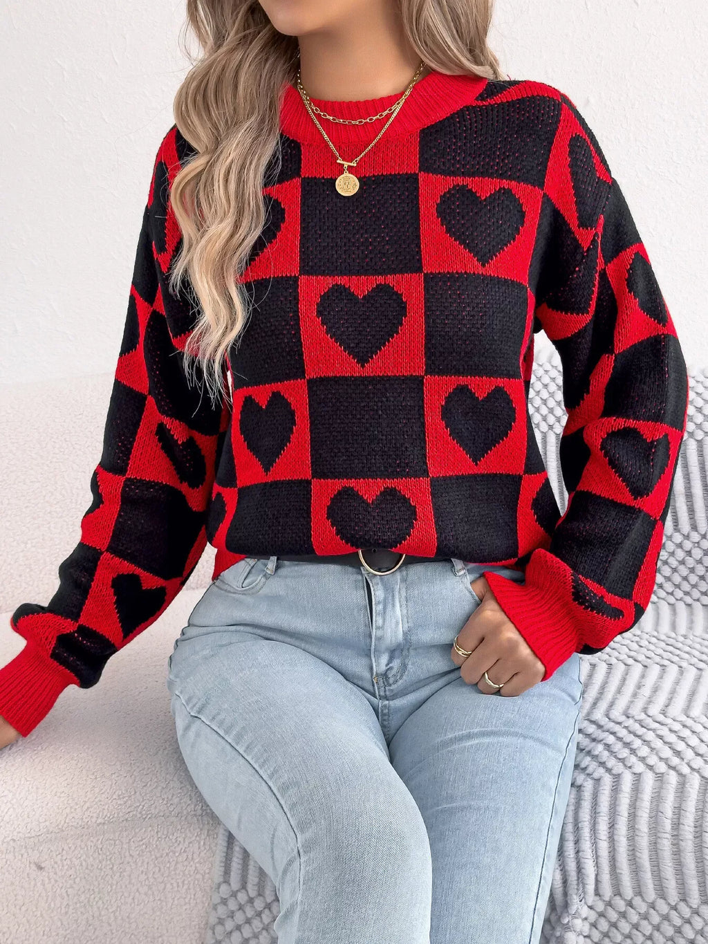 Woman wearing a red and black checkerboard knit sweater with black heart patterns and light wash denim jeans.